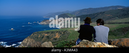 Two men sitting on rock overlooking the Pacific Ocean coastline view south of Big Sur California with Bixby Bridge and Big Creek Stock Photo