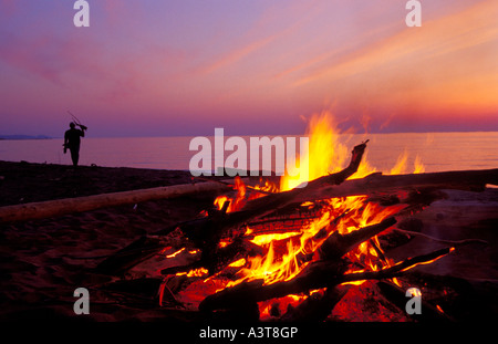 A CAMPFIRE BURNS ON A LAKE SUPERIOR BEACH AT DUSK IN ONTONAGON MICHIGAN AS A SILHOUETTED MAN CARRIES DRIFTWOOD Stock Photo