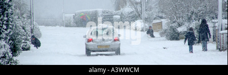 Village road with snow falling kids playing people walking and car driving along with rear lights on Stock Photo