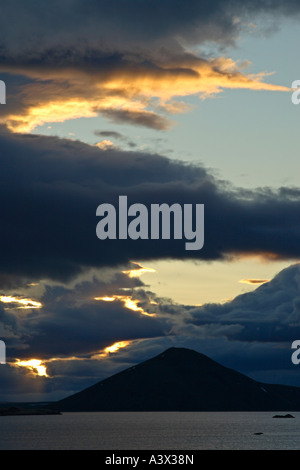 View across Myvatn at dusk towards a rootless cone hill clouds rim lit by setting sun Iceland Stock Photo