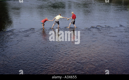 three people crossing a river via stepping stones one person is gesturing to another for help Bolton Abbey in Yorkshire across the River Wharfe Stock Photo