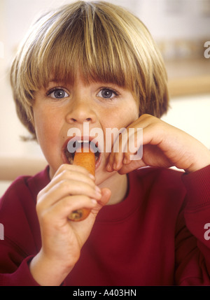Young boy sitting in a kitchen taking a bite out of a carrot Stock Photo