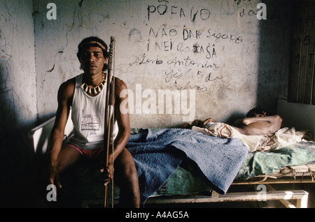 Aruana Pataxo sits on his father's bed.  A rancher's gunmen  arrived to chase the Pataxo from their land. The old man confronted them with his bow arrow. they shot him with heavy calibre rifles and he has been bedridden ever since. Stock Photo