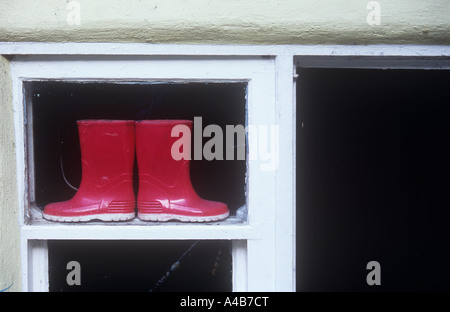 Close up of pair of childs red wellington boots sitting back-to-back on ledge of small window and viewed from outside Stock Photo