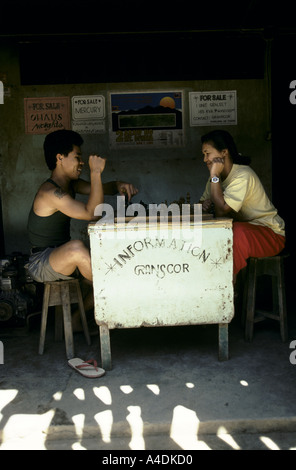 A gold dealer plays chess with a woman in the shade under various  "For Sale" signs. Mount Diwata, Mindanao, The Philippines Stock Photo