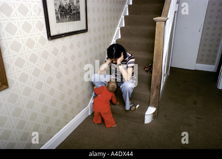 Woman suffering from depression sitting on a step at the bottom of the stairs with child looking at her Stock Photo