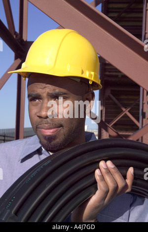 African ethnicity construction worker wearing yellow hardhat and carrying coiled tubing outdoors. Stock Photo