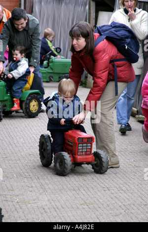 Toddler riding a small red toy tractor whilst mother in red jacket with backpack holds him Stock Photo