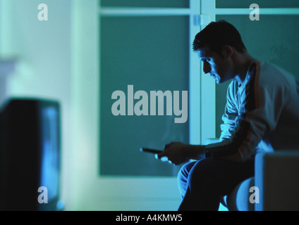 Man sitting on sofa watching television, remote control in hand, side view Stock Photo