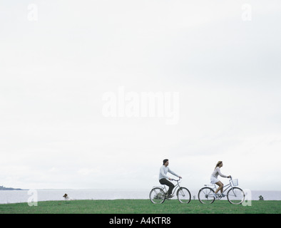Young man and woman bicycling on grass with sea and overcast sky in background Stock Photo