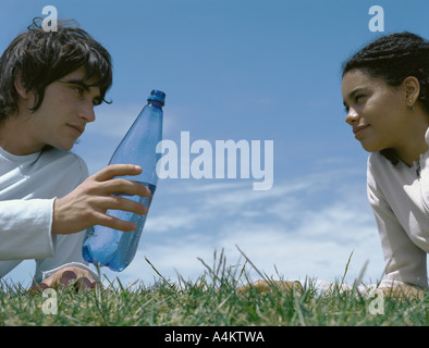 Young man and young woman leaning on elbows in grass, young man holding out bottle Stock Photo