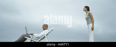 Man sitting in lounge chair with laptop on lap, woman standing peering over man's shoulder, side view Stock Photo