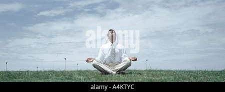 Man in business clothes sitting in lotus position on grass with head back and eyes closed, front view Stock Photo
