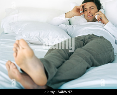 Clothed man lying on bed talking on cordless phone, hand to head Stock Photo