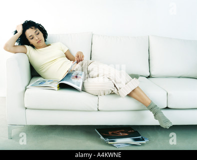 Woman lounging on sofa looking at magazine with stack of magazines on floor, full length Stock Photo