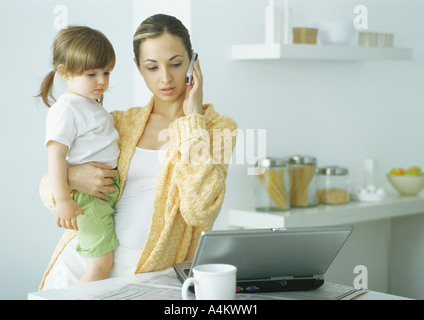 Young woman holding little girl with one arm, holding cell phone to ear with other hand, looking down at laptop Stock Photo