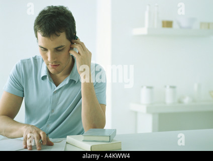 Young man in polo shirt sitting at table with books and papers, holding cell phone, looking down Stock Photo