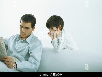 Man sitting on sofa reading newspaper, boy standing behind sofa looking over man's shoulder Stock Photo