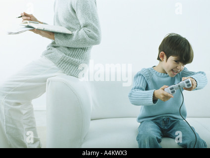 Man sitting on arm of sofa writing in agenda, boy sitting on sofa with joystick Stock Photo