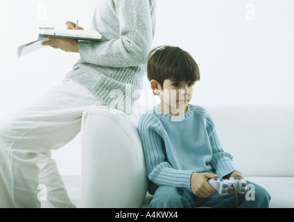 Man sitting on arm of sofa writing in agenda, boy sitting on sofa with joy stick Stock Photo