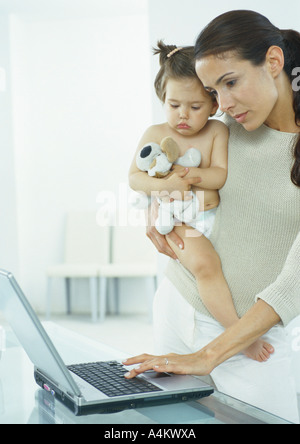 Woman holding little girl in one arm, typing on laptop with other hand Stock Photo