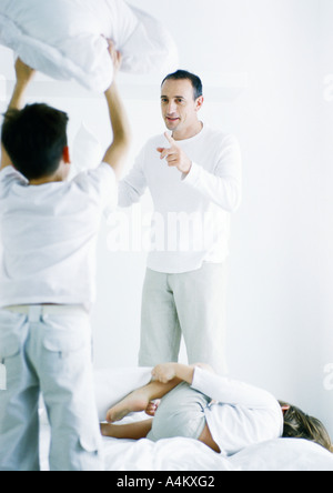 Boy standing on bed holding up pillow, rear view, second boy lying on bed, man pointing at first boy Stock Photo