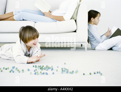 Boy playing marbles on floor, woman and girl reading in background
