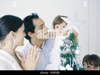 Little girl in fathers arms touching star on Christmas tree next to mother and brother Stock Photo