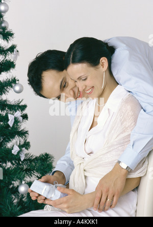 Woman holding gift, man bending over her shoulder, Christmas tree in background Stock Photo