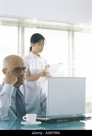 Businessman sitting at laptop with cup and saucer, woman standing behind him taking notes Stock Photo