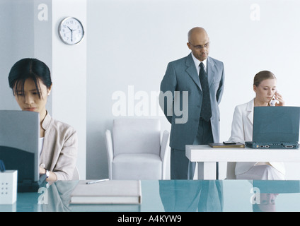 Two women working on laptops in office, supervisor looking over one woman's shoulder Stock Photo