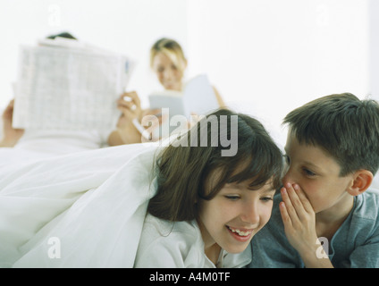 Boy whispering to girl, parents reading in background Stock Photo