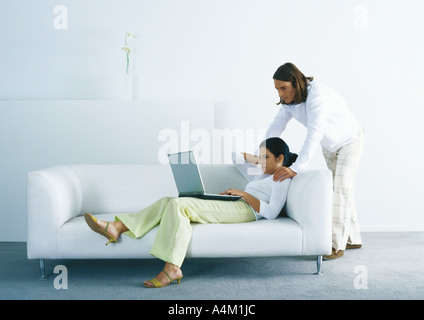 Woman lounging on sofa and using laptop, man standing, looking over woman's shoulder