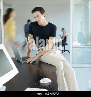 Man sitting on edge of desk in busy office Stock Photo