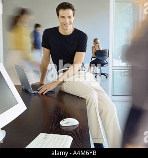 Man sitting on edge of desk in busy office Stock Photo
