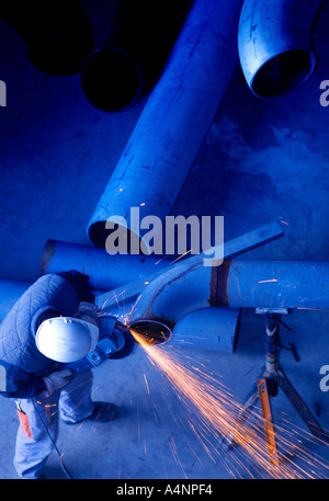 Man wearing an white hard hat in a factory working on a gas pipeline tube using an angle grinder Stock Photo