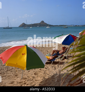 Pair of two colourful sun umbrellas with man and woman sun tanning on sandy Reduit Beach St Lucia The Caribbean Stock Photo