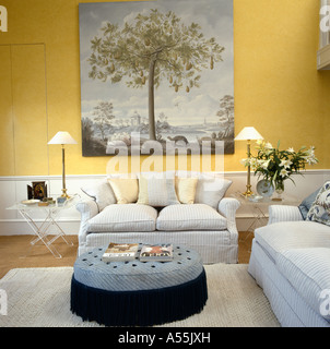 Large 18th-century style picture above pale gray sofa in pale yellow living room with large circular upholstered stool Stock Photo