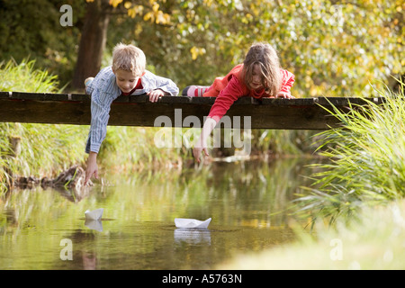 Boy (10-13) and girl (7-9) lying on bridge watching paper boats in water Stock Photo