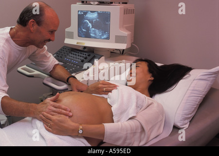 Proud husband feeling abdomen of pregnant wife during ultrasound test with fetus on screen Stock Photo