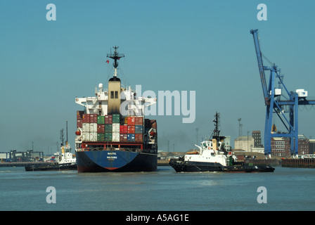 Arriving cargo container freighter ship stacked full with load of shipping containers tugboats push & pull towards cranes Felixstowe port Suffolk UK Stock Photo