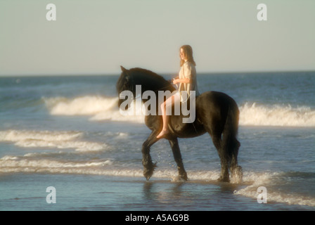 Young woman riding Friesian stallion on beach at sunset Stock Photo