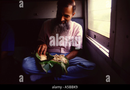 An Indian commuter sits crossed legs and eats South Indian Thali meal on banana leaf with his hands in a train Tamil Nadu state South India Stock Photo