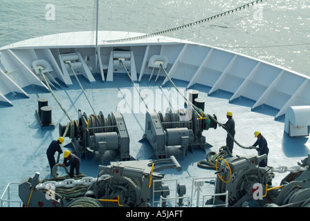 Looking down view from above at bow of cruise ship liner teamwork of seamen in hard hats handling mooring ropes & working winding gear machines Stock Photo