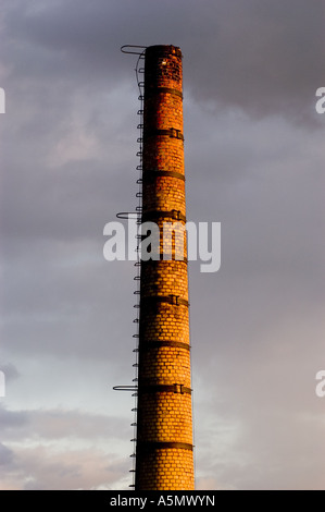 Old brick long closed chimney standing tall against a dramatic cloudy sky, featuring a metal ladder and reinforcing bands. Stock Photo