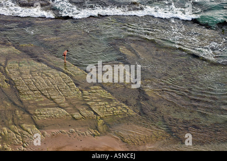 Shore Exploration: A man walks on the channeled rocky shore below the stair. A wave is washing in from the deeper water Stock Photo