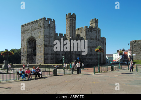 Tourists & locals on blue sky day in Welsh Castle Square with historical medieval stone fortress of Caernarfon castle beyond Gwynedd North Wales UK Stock Photo