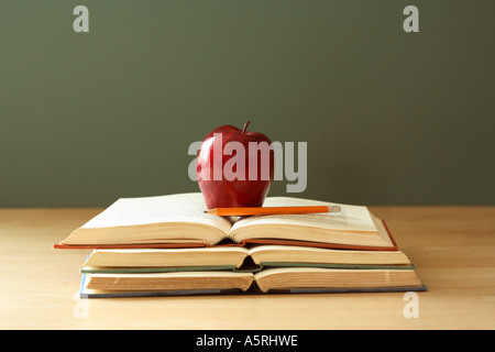 Stack of open school books, red apple and pencil on desk with chalkboard in background. Stock Photo