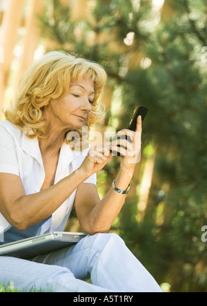 Senior woman using cell phone, laptop on lap, outdoors Stock Photo