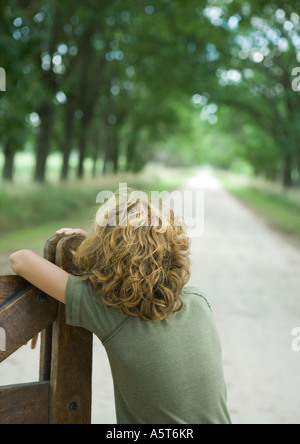 Boy leaning on gate, facing dirt road, rear view Stock Photo